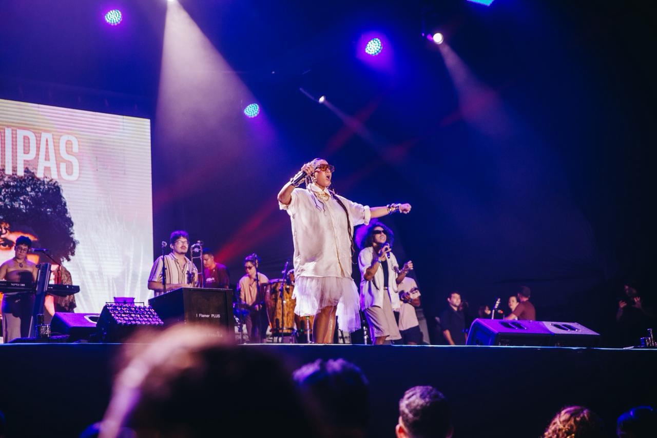 Cantora com roupa branca e braços estendidos performa em palco iluminado por luzes azuis e roxas, com banda ao fundo.