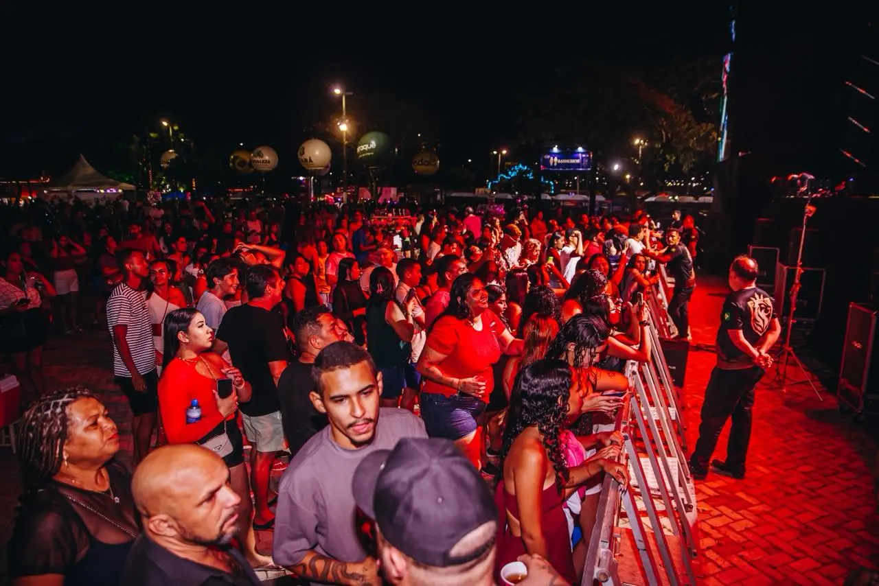 Multidão reunida à noite em frente ao palco de um show ao ar livre, com iluminação vermelha, público animado e seguranças posicionados na área frontal do evento.