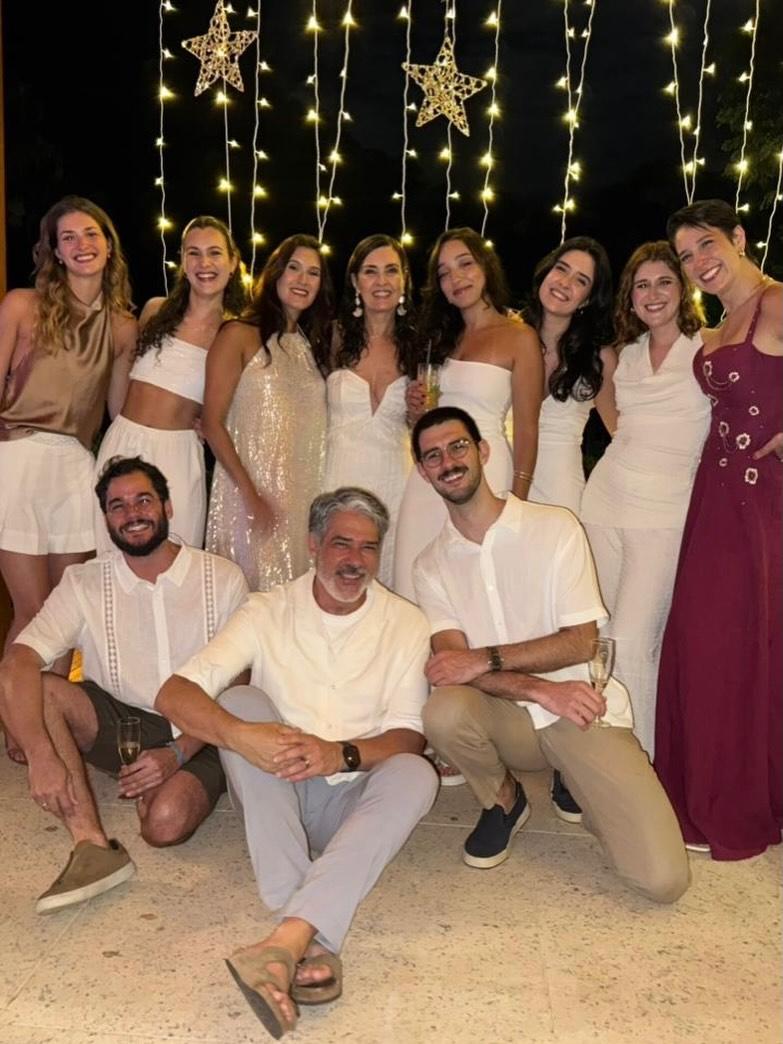Na imagem, uma fotografia de grupo num ambiente festivo noturno, capturando um momento de celebração. O grupo é composto por onze pessoas, sendo oito mulheres de pé ao fundo e três homens agachados ou sentados à frente. Todos sorriem para a câmara. No centro do grupo de pé, está a jornalista Fátima Bernardes, que veste um vestido branco com decote em 