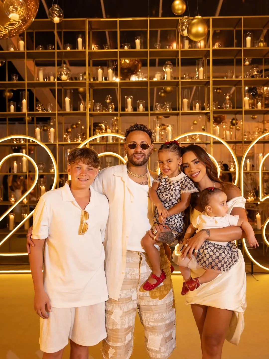 Na imagem, fotografia da família de Neymar Jr. posando em um evento festivo iluminado. Da esquerda para a direita, estão o filho mais velho Davi Lucca, o jogador Neymar Jr. segurando a filha Mavie no colo, e Bruna Biancardi segurando a filha caçula, Helena. Todos vestem roupas em tons de branco e bege. As duas crianças usam conjuntos com estampas de logotipos de grife e sapatinhos vermelhos idênticos. Neymar usa óculos escuros e correntes douradas. O fundo apresenta uma estante dourada repleta de velas acesas e luzes neon amarelas que formam o número 