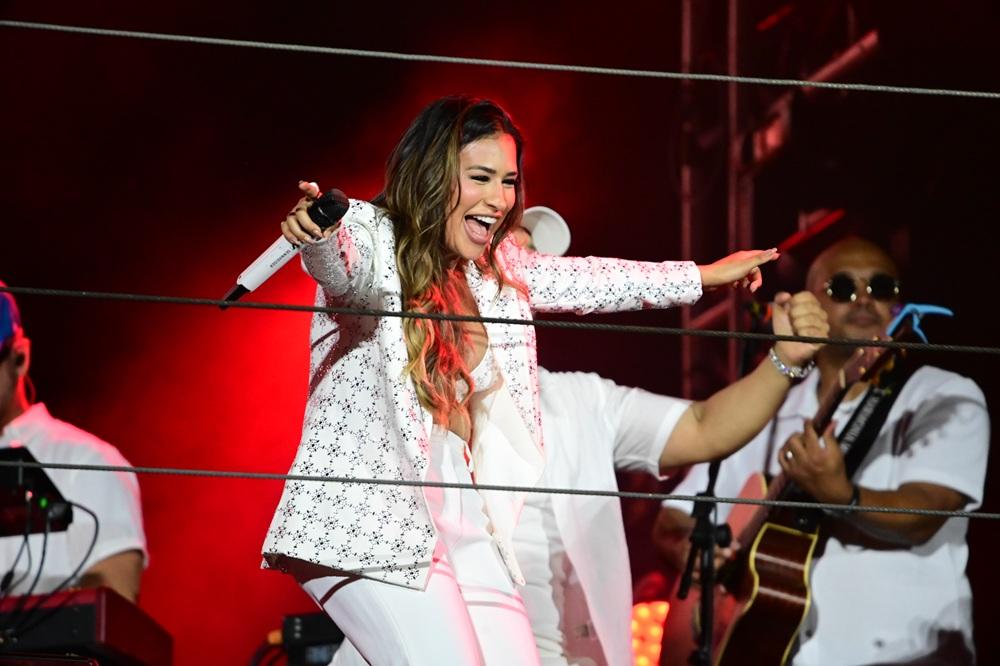 Na imagem, fotografia da cantora Simone Mendes cantando alegremente em um palco. A cantora é capturada de perfil, sorrindo abertamente com o microfone na mão direita e o braço esquerdo estendido. Ela tem cabelos castanhos longos e ondulados. Veste um conjunto branco composto por um blazer com pequenos bordados escuros e calças combinando. O ambiente é escuro com iluminação avermelhada ao fundo. Cabos de aço horizontais atravessam a frente da imagem, e parte da banda é visível atrás dela.