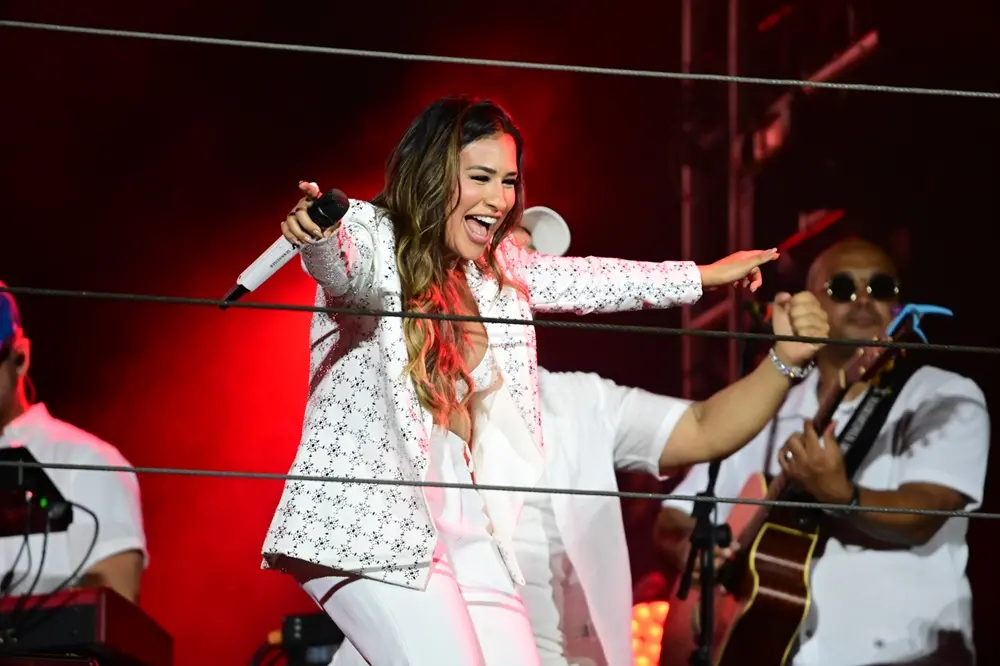 Na imagem, fotografia da cantora Simone Mendes cantando alegremente em um palco. A cantora é capturada de perfil, sorrindo abertamente com o microfone na mão direita e o braço esquerdo estendido. Ela tem cabelos castanhos longos e ondulados. Veste um conjunto branco composto por um blazer com pequenos bordados escuros e calças combinando. O ambiente é escuro com iluminação avermelhada ao fundo. Cabos de aço horizontais atravessam a frente da imagem, e parte da banda é visível atrás dela.