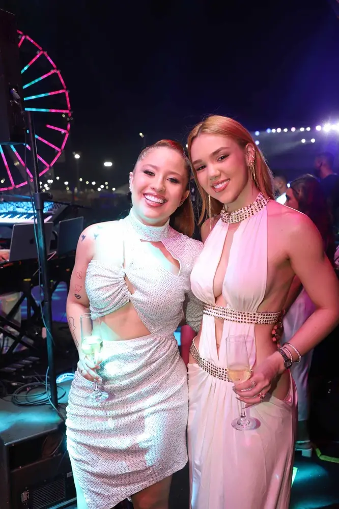 Na imagem, fotografia das influenciadoras Mari Fernandez e Julia Ribeiro posando juntas em um evento noturno. As duas aparecem sorridentes, abraçadas, cada uma segurando uma taça de bebida. Ambas usam vestidos brancos com recortes e detalhes brilhantes ou metálicos. O ambiente de fundo é festivo, com luzes coloridas e uma estrutura que lembra uma roda-gigante iluminada à esquerda.