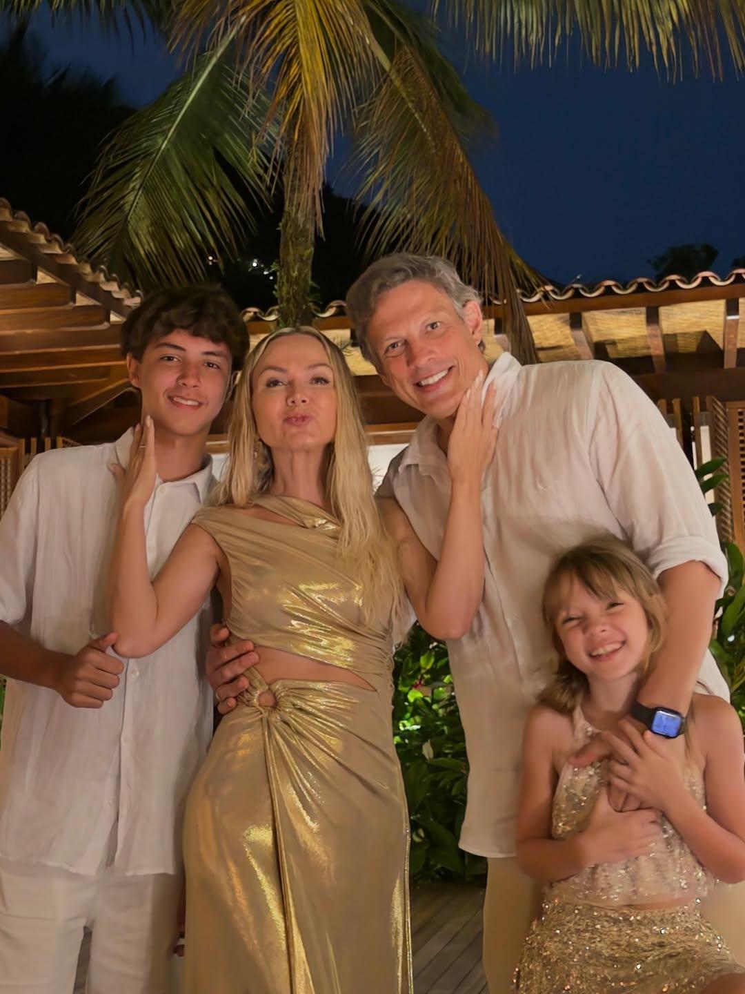 Na imagem, fotografia da apresentadora Eliana com seu marido, Adriano Ricco, e seus dois filhos em um ambiente tropical. Eliana está ao centro fazendo um biquinho para a foto, ladeada pelo filho mais velho à esquerda e pelo marido à direita, que segura a filha caçula à frente. Eliana usa um vestido dourado metalizado com recortes na cintura. A filha usa um vestido de paetês dourados e os homens vestem camisas brancas. O grupo está sob uma estrutura de madeira com telhado aparente, tendo uma palmeira e o céu noturno ao fundo.