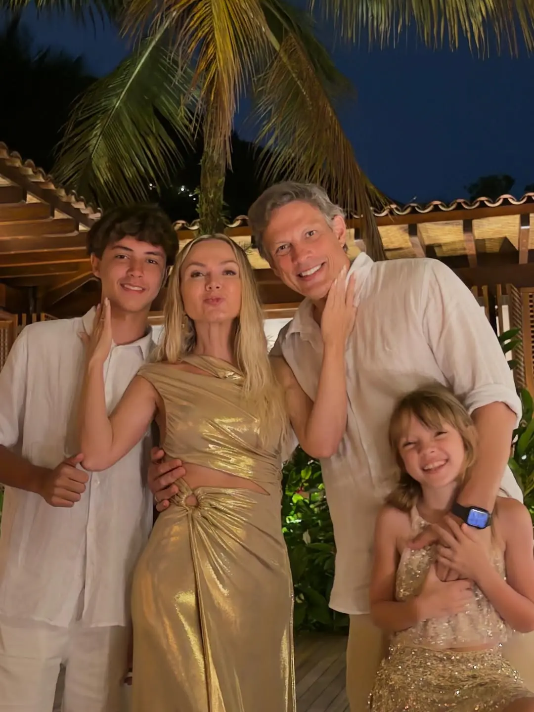 Na imagem, fotografia da apresentadora Eliana com seu marido, Adriano Ricco, e seus dois filhos em um ambiente tropical. Eliana está ao centro fazendo um biquinho para a foto, ladeada pelo filho mais velho à esquerda e pelo marido à direita, que segura a filha caçula à frente. Eliana usa um vestido dourado metalizado com recortes na cintura. A filha usa um vestido de paetês dourados e os homens vestem camisas brancas. O grupo está sob uma estrutura de madeira com telhado aparente, tendo uma palmeira e o céu noturno ao fundo.