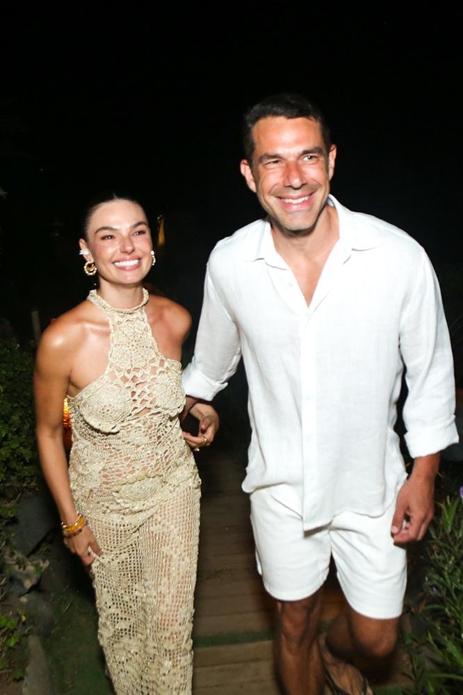 Na imagem, O casal caminha sorridente em um ambiente externo à noite. Isis Valverde usa um vestido de crochê bege com gola alta e Marcos Buaiz veste camisa e bermuda brancas.