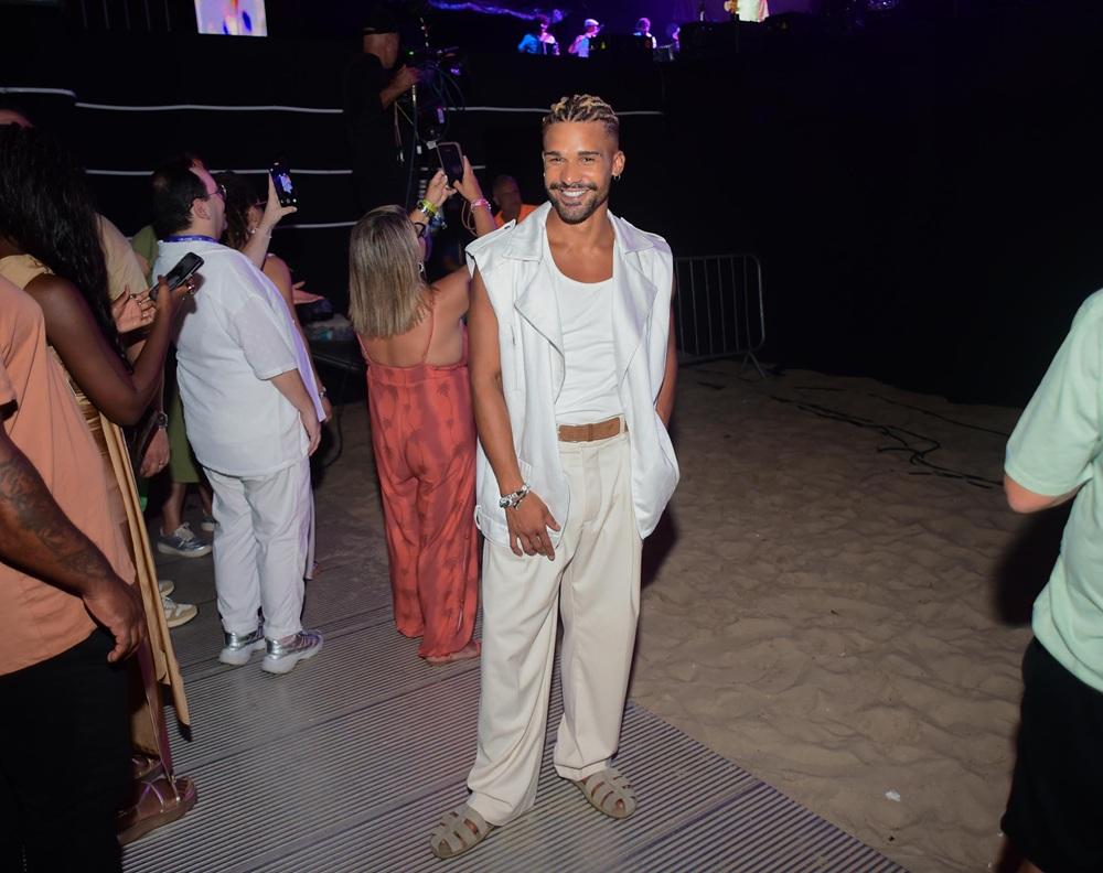 Na imagem, o ator Lucas Leto sorri para a câmera em um evento noturno na praia. Ele veste um look todo em tons de branco e bege: colete estruturado sem mangas, regata branca, calças de alfaiataria largas e sandálias estilo 