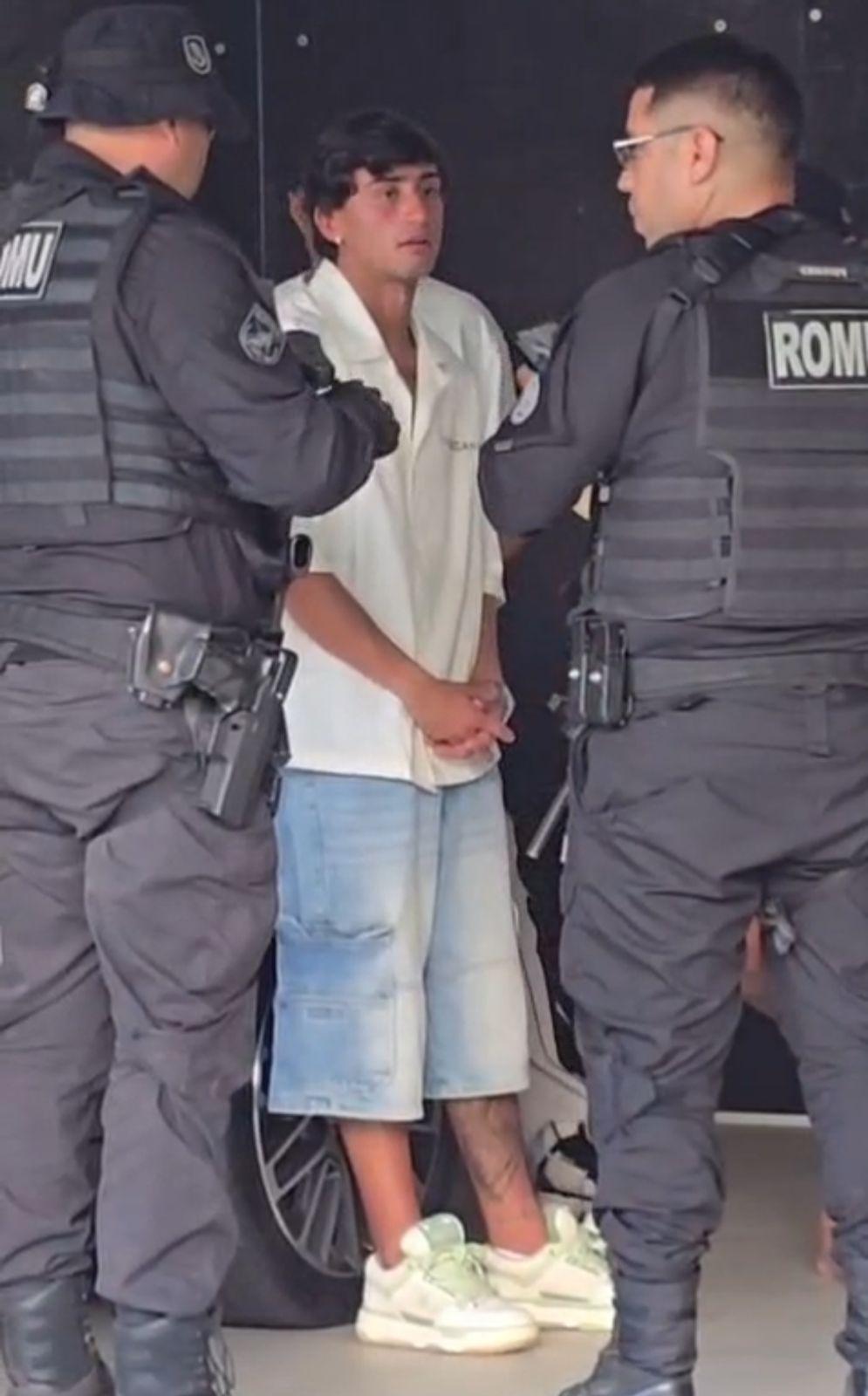 O jogador Herrera, vestindo camisa branca e bermuda jeans, aparece sob custódia de dois agentes da Guarda Municipal fardados e armados. Ele mantém as mãos cruzadas à frente do corpo com uma expressão séria, em uma composição que foca na abordagem policial em um ambiente urbano.