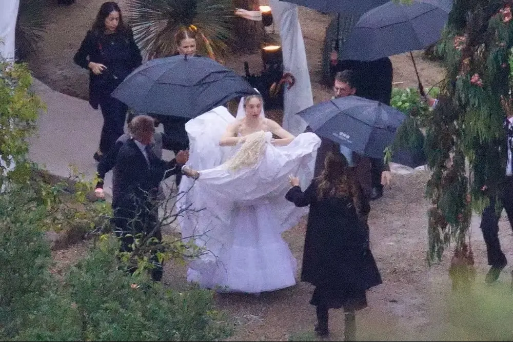 Este Haim levantando seu vestido branco de noiva, ao caminhar, rodeada de pessoas com guarda-chuvas preto.