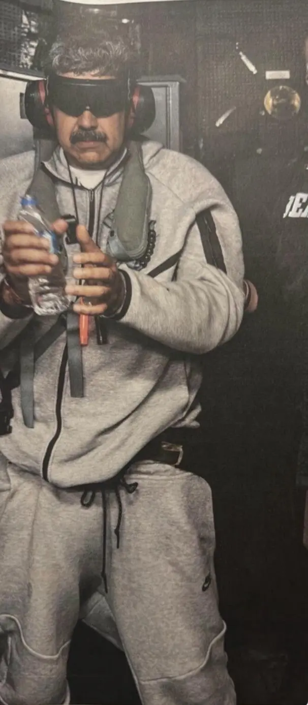 Na imagem, um homem de pele clara e bigode escuro espesso (semelhante ao presidente venezuelano Nicolás Maduro) em um ambiente fechado e escuro. Ele veste um conjunto de moletom cinza da Nike (modelo Tech Fleece) e segura uma garrafa de água plástica. O que chama a atenção é o equipamento de segurança que ele utiliza: protetores auriculares robustos (pretos com detalhes vermelhos) e óculos de proteção totalmente escurecidos, que cobrem seus olhos como uma viseira. Sobre o moletom, ele usa um colete de segurança ou flutuador cinza com tiras pretas e um apito laranja pendurado. A iluminação é dramática, focada no homem, enquanto o fundo permanece em sombras, sugerindo o interior de uma aeronave militar ou um ambiente industrial.