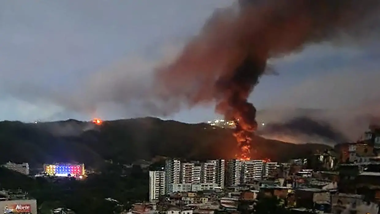 Na imagem, fotografia noturna ou ao entardecer de uma encosta urbana na Venezuela, mostrando um grande incêndio. Uma densa coluna de fumaça escura e avermelhada sobe verticalmente de um ponto nas colinas, dominando o céu nublado. Abaixo da fumaça, chamas intensas são visíveis. Em primeiro plano, há prédios residenciais altos e aglomerados de casas menores (comunidades) construídas no morro. À esquerda, as luzes da cidade brilham, incluindo um edifício com iluminação colorida, criando um forte contraste entre a vida urbana cotidiana e a emergência do incêndio ao fundo.