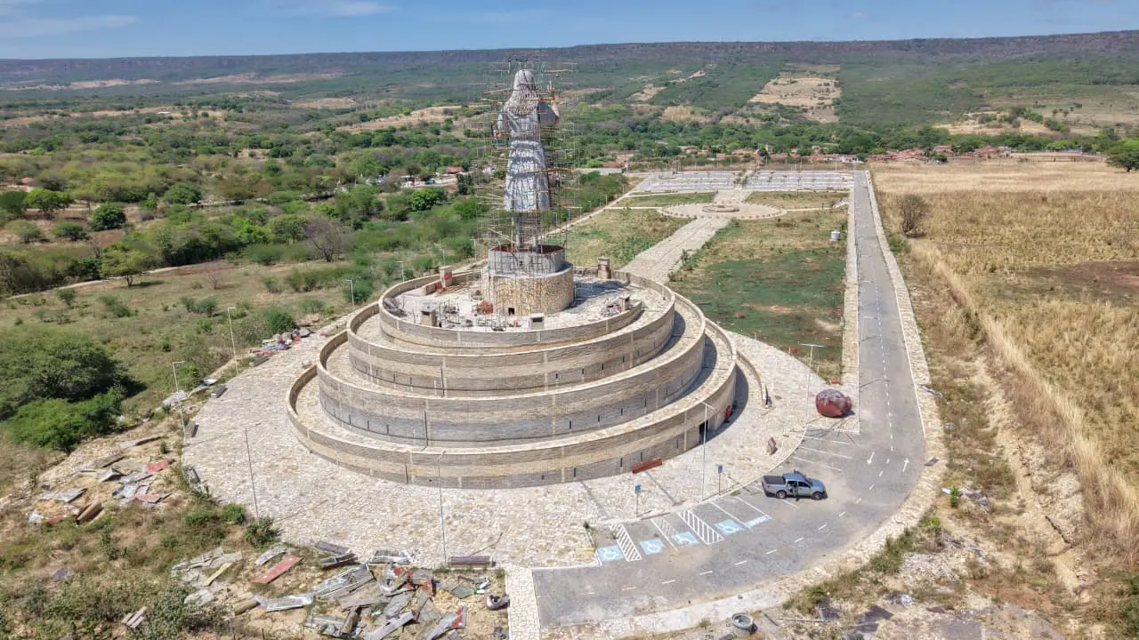 Vista aérea do Complexo da Menina Benigna em construção, destacando a grande estátua cercada por andaimes sobre uma base circular de múltiplos níveis, situada em uma área rural com vegetação rasteira e montanhas ao fundo.
