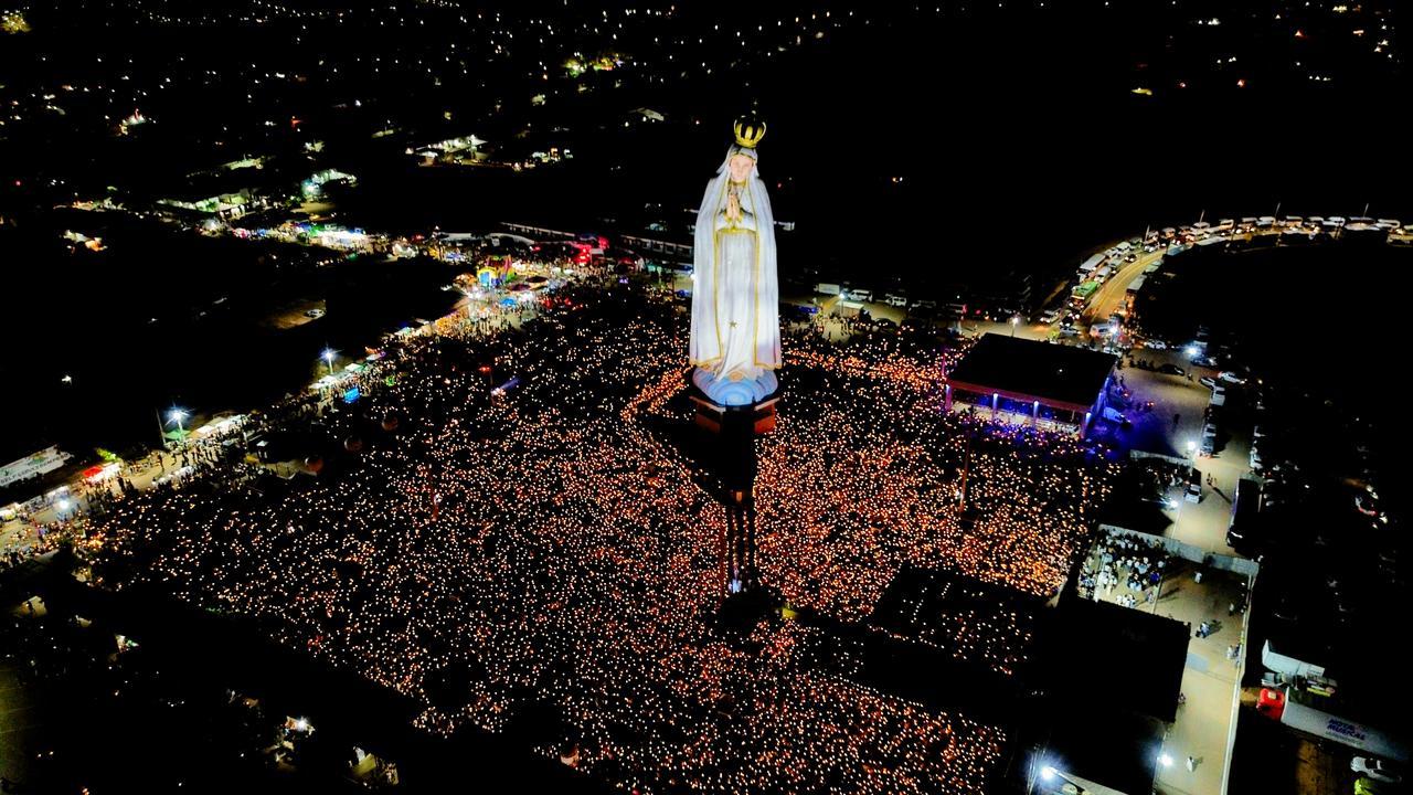 Vista aérea noturna da estátua de Nossa Senhora de Fátima em Crato, Ceará, cercada por uma vasta multidão de fiéis segurando velas durante uma celebração religiosa.