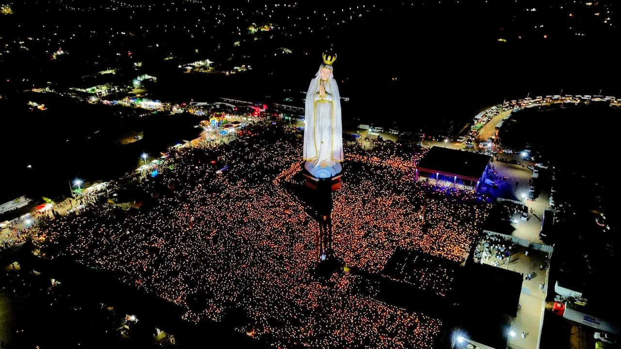 Vista aérea noturna da estátua de Nossa Senhora de Fátima em Crato, Ceará, cercada por uma vasta multidão de fiéis segurando velas durante uma celebração religiosa.