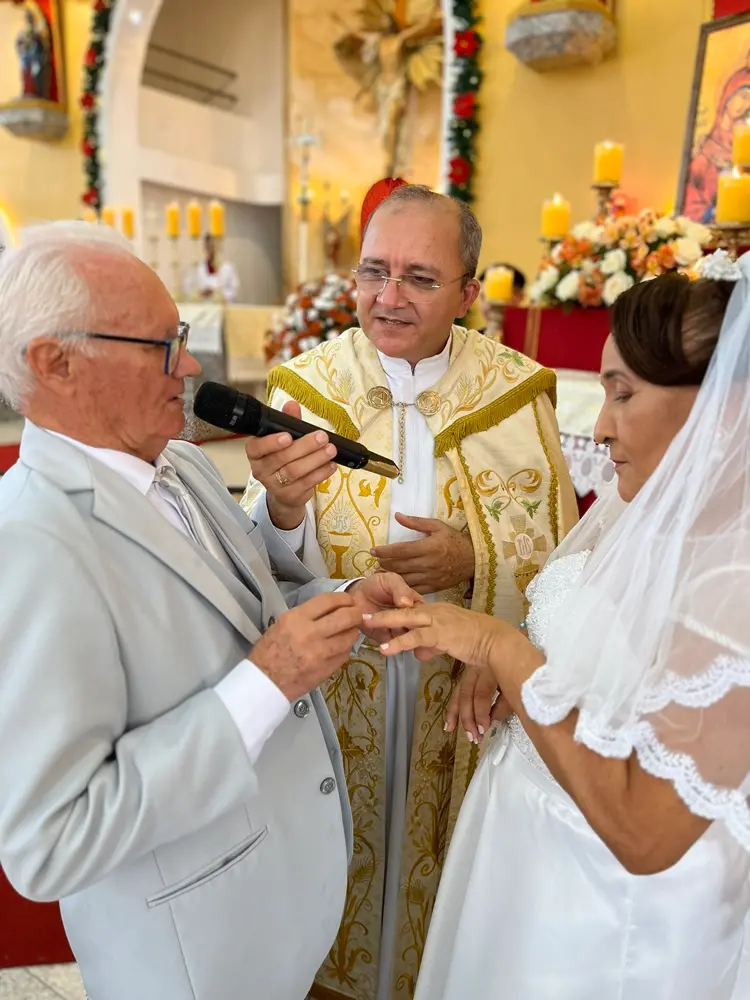 Na imagem, uma fotografia em close de uma cerimônia de casamento dentro de uma igreja católica. No centro, um padre de óculos e vestimenta litúrgica branca com bordados dourados segura um microfone para um homem idoso à esquerda. O homem, que tem cabelos brancos e usa um terno cinza claro, está colocando uma aliança no dedo de uma mulher idosa à direita. A noiva veste um vestido branco e um véu com detalhes em renda. O fundo mostra o altar da igreja decorado com flores brancas e laranjas, velas acesas e elementos sacros.