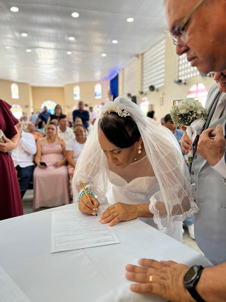 Na imagem, uma noiva idosa, em primeiro plano e levemente inclinada, assina um documento matrimonial sobre uma mesa coberta com uma toalha branca. Ela usa um vestido de noiva branco com detalhes em pérolas, um véu translúcido com bordas de renda e uma tiara de pérolas. À sua direita, parte do noivo é visível, vestindo um terno cinza e segurando um pequeno buquê de flores brancas. Ao fundo, o interior de uma igreja com convidados sentados em bancos de madeira sob uma iluminação suave. A composição foca no momento solene da assinatura.