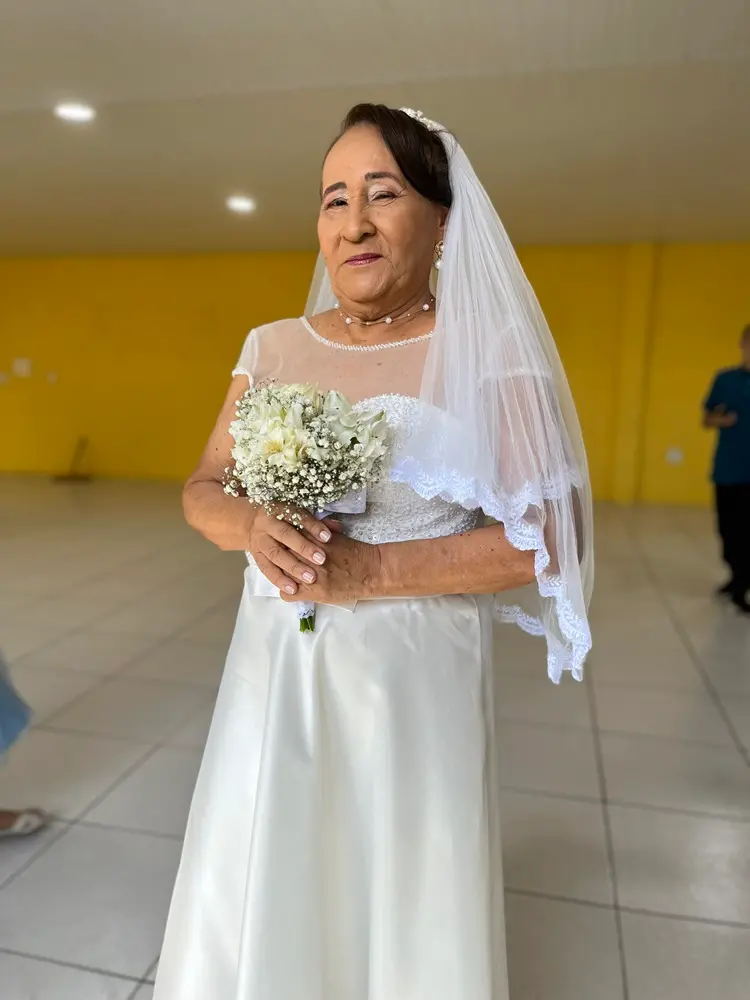Na imagem, retrato de corpo inteiro de uma noiva idosa posando em um salão com piso claro e parede amarela ao fundo. Ela sorri suavemente enquanto segura um buquê de flores brancas e miçangas (mosquitinhos) com as duas mãos. Veste um vestido de noiva branco acetinado com detalhes em renda no decote e um véu translúcido longo. A iluminação é frontal e destaca os detalhes das joias de pérola que ela usa.