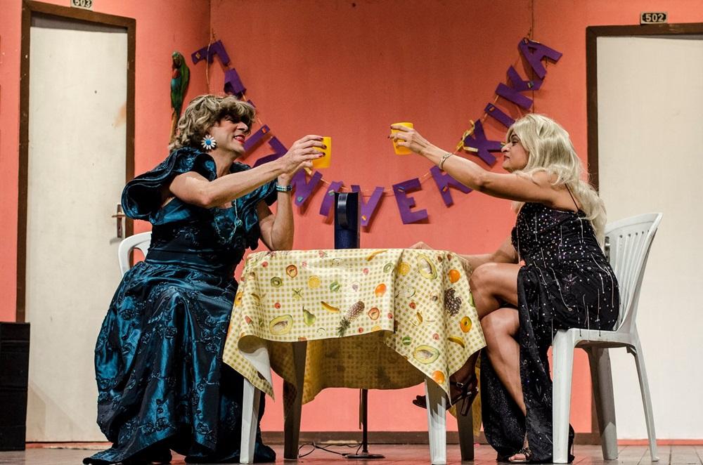 Na imagem, fotografia de uma cena teatral com duas pessoas caracterizadas como mulheres, sentadas à mesa e brindando com copos amarelos. A pessoa à esquerda usa um vestido longo azul brilhante e peruca castanha; a da direita usa um vestido preto de fenda e peruca loira. Elas estão em um cenário simples com paredes salmão, duas portas brancas ao fundo e uma faixa roxa pendurada onde se lê 