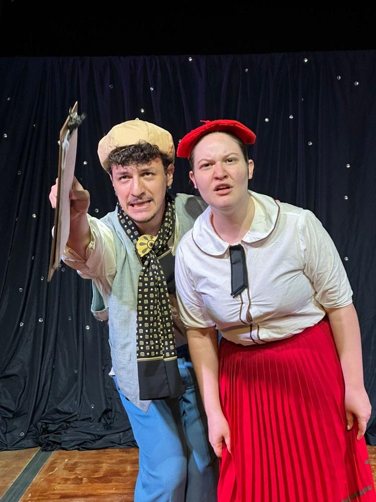 Na imagem, fotografia em plano médio de dois atores em uma performance teatral. À esquerda, um homem com expressão carrancuda segura uma prancheta e aponta para frente; ele usa boina bege, colete cinza, calças azuis e um cachecol preto com estampas douradas. À direita, uma mulher com expressão de confusão ou desdém olha para a mesma direção; ela veste uma boina vermelha, blusa branca de gola arredondada e uma saia longa vermelha plissada. O fundo é composto por uma cortina preta com pequenos pontos brilhantes que simulam estrelas, sobre um palco de madeira.