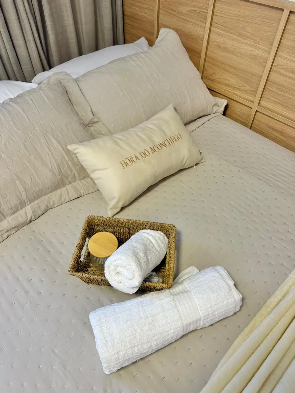 Na imagem, close-up de uma cama de casal arrumada com enxoval em tons de bege e fendi. Sobre a colcha texturizada, há uma almofada decorativa com a frase bordada 'HORA DO ACONCHEGO'. À frente, uma cesta de palha rústica contém uma toalha branca enrolada e um frasco com tampa de madeira, acompanhada de outra toalha branca maior também enrolada. Ao fundo, vê-se parte de uma cabeceira em madeira clara com design ripado e cortinas neutras, criando uma atmosfera serena e acolhedora.