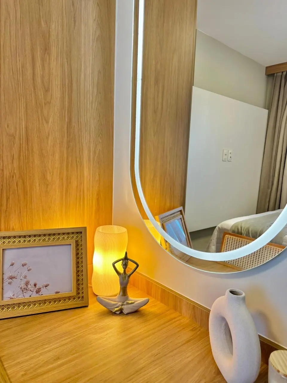 Na imagem, close de um canto decorativo com móveis em madeira clara. Um espelho oval com moldura de LED iluminada reflete parte do quarto. Sobre a bancada, encontram-se itens decorativos: um porta-retrato com moldura de palha sextavada, uma luminária de mesa ondulada com luz quente, uma pequena escultura de uma pessoa em posição de yoga e um vaso de cerâmica bege com design orgânico vazado. A cena transmite uma sensação de tranquilidade e sofisticação.