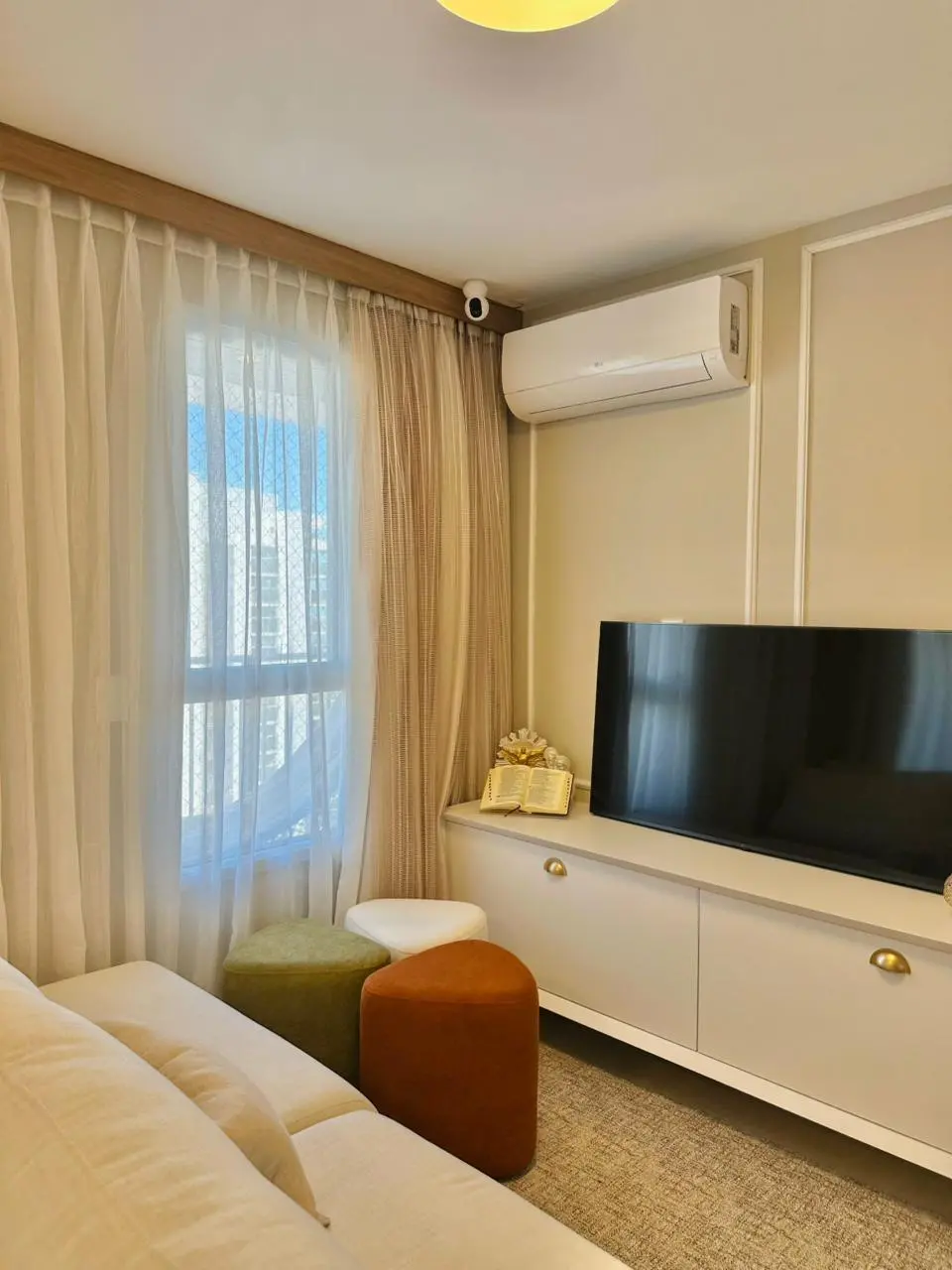Na imagem, ambiente de estar integrado com decoração contemporânea. À esquerda, um sofá de tecido claro com almofadas. À frente, um rack branco com puxadores dourados suporta uma televisão de tela plana. Sobre o rack, um livro aberto e adornos decorativos. No chão, três puffs em formatos orgânicos e cores terrosas (verde musgo, terracota e branco) trazem um toque de cor sobre o tapete cinza. Uma grande janela com cortinas de tecido fluido e translúcido permite a entrada de luz natural, e um ar-condicionado branco está instalado acima do móvel.