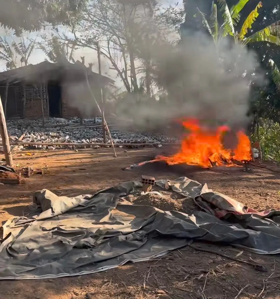 Queima e maconha uma área rural com fogo e fumaça, casas de madeira ao fundo e vegetação ao redor, indicando uma emergência ou acidente.