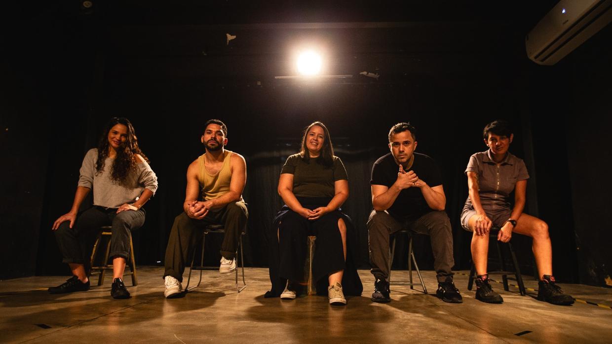 Na imagem, cinco pessoas, três mulheres e dois homens, estão sentadas lado a lado em bancos em um palco de teatro escurecido. Elas olham frontalmente para a câmera com expressões serenas. O ambiente é iluminado por um único refletor central ao fundo, que cria um efeito de auréola acima do grupo e destaca as texturas do chão de cimento polido. Da esquerda para a direita: uma mulher de cabelos longos e cacheados em uma blusa cinza; um homem de regata bege; uma mulher ao centro com camiseta verde oliva e calça escura; um homem de camiseta preta com as mãos entrelaçadas; e uma mulher de cabelos curtos em um macacão cinza. O clima é intimista e artístico.