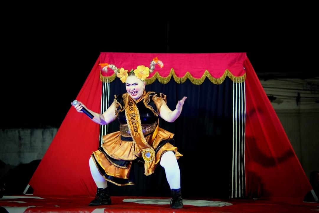 Na imagem, uma artista performática está no centro de um palco de circo, em uma pose expressiva e agachada. Ela tem a pele branca, usa maquiagem artística carregada com sobrancelhas pretas marcadas e um sorriso vibrante. Seu cabelo está preso em dois rabos de cavalo altos e esculpidos para cima, adornados com grandes flores amarelas e laranjas. Ela veste um figurino de estilo circense nas cores dourado e preto, com saia de babados, meias brancas e botas pretas. Atravessada no peito, usa uma faixa dourada com a inscrição 