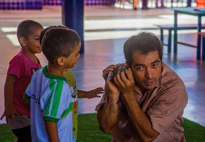 Na imagem, um homem adulto está agachado em um gramado sintético verde, interagindo com duas crianças pequenas em um ambiente escolar ou comunitário. O homem segura um pequeno objeto artesanal (como um chocalho de tecido) próximo ao ouvido, com uma expressão de escuta atenta e curiosidade. À sua frente, um menino com camiseta branca e listras verdes sorri, observando a ação. Ao fundo, outra criança com camiseta rosa observa a cena. O ambiente é iluminado e possui colunas azuis e mesas de madeira ao fundo.