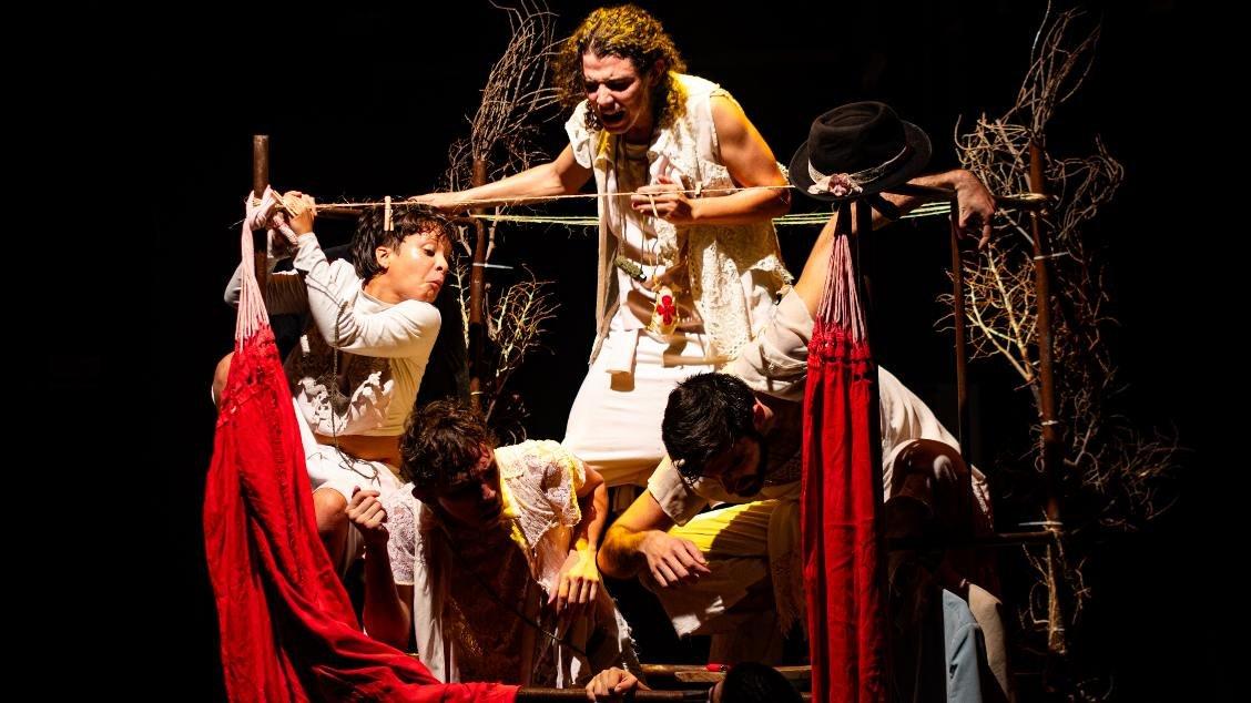 Na imagem, uma fotografia de uma cena teatral do espetáculo 'Um pé de quê', do Grupo Lindo. No centro, quatro atores com expressões intensas e dramáticas ocupam uma estrutura de madeira que remete a uma balsa ou cercado rústico. À esquerda, uma mulher de branco puxa uma corda com esforço. No topo, outra mulher grita fervorosamente. Abaixo, dois homens completam a composição, um deles agachado. O cenário é composto por galhos secos e dois tecidos vermelhos vibrantes que pendem das laterais, assemelhando-se a redes. A iluminação é focada nos atores contra um fundo totalmente preto, criando um forte contraste dramático.