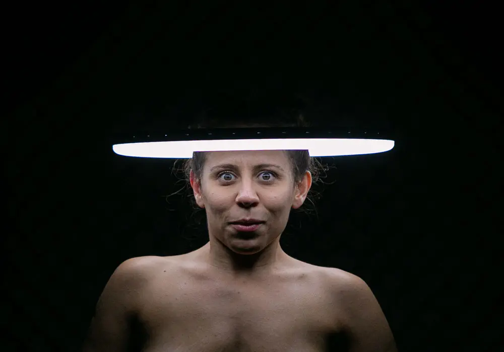 Na imagem, fotografia em close de uma mulher com expressão de espanto sob um fundo preto. Um anel de luz branco e brilhante está posicionado horizontalmente à altura de sua testa, criando um efeito de auréola tecnológica e iluminando intensamente seus olhos arregalados.