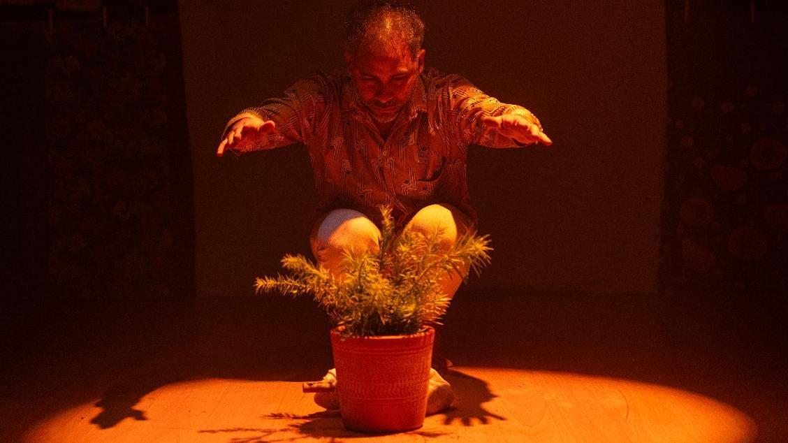 Na imagem, um homem agachado em um palco escuro, sob uma luz cenográfica âmbar intensa que incide de cima para baixo. Ele estende as mãos sobre um vaso de planta no chão, como se realizasse um ritual ou gesto de cuidado. A iluminação cria sombras marcantes e destaca a planta no centro da cena.