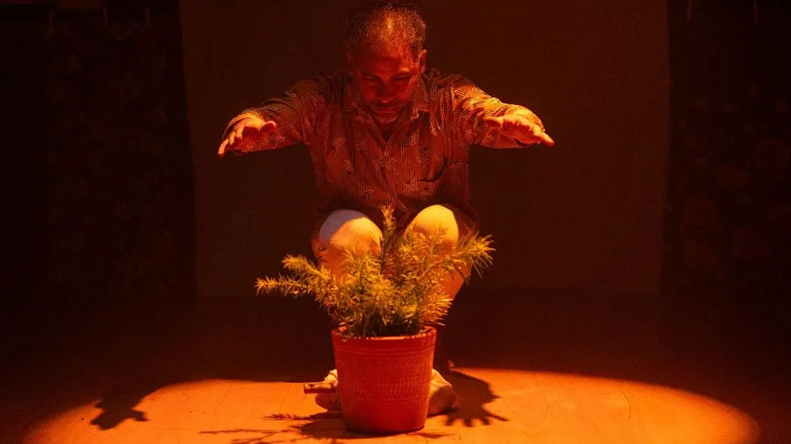 Na imagem, um homem agachado em um palco escuro, sob uma luz cenográfica âmbar intensa que incide de cima para baixo. Ele estende as mãos sobre um vaso de planta no chão, como se realizasse um ritual ou gesto de cuidado. A iluminação cria sombras marcantes e destaca a planta no centro da cena.