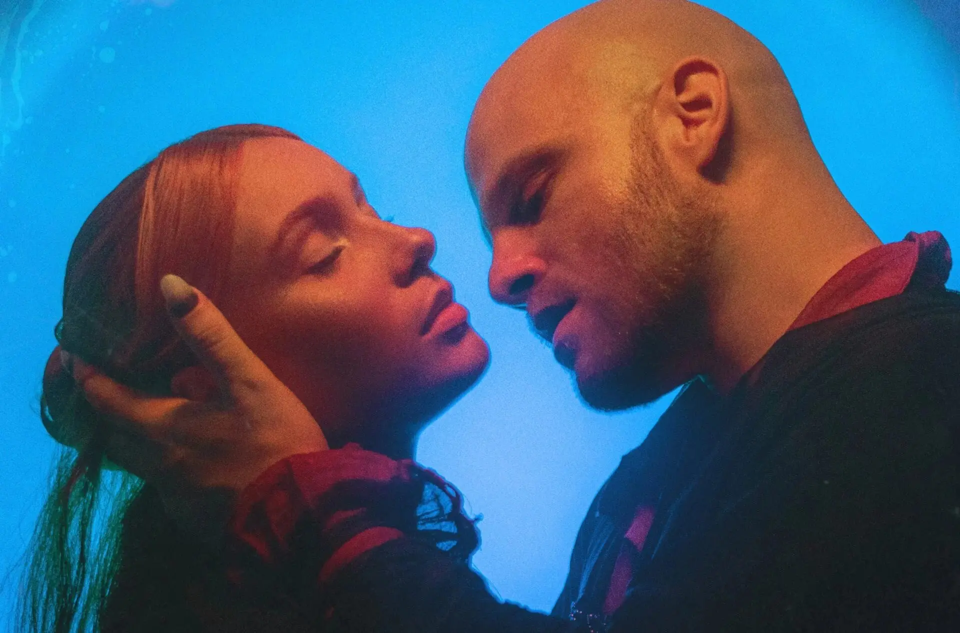 Na imagem, close-up de dois atores em um momento íntimo e dramático. Uma mulher de cabelos ruivos está com os olhos fechados enquanto um homem careca aproxima o rosto do dela, segurando sua cabeça com delicadeza. A cena é banhada por uma luz azul vibrante ao fundo, que contrasta com a iluminação quente nos rostos.