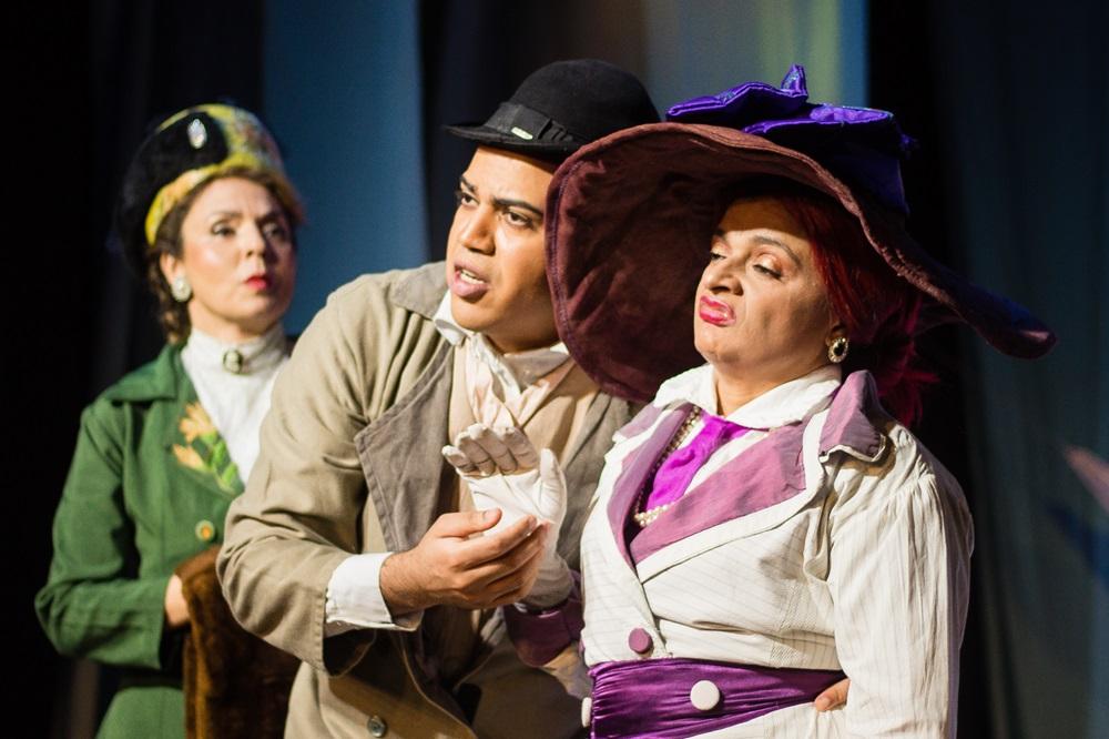 Na imagem, três atores em cena de uma peça de teatro, vestindo figurinos de época inspirados no início do século XX. No centro, um homem jovem com expressão de urgência fala com uma mulher à direita. Ela usa um chapéu roxo desproporcionalmente grande, tem uma expressão de desdém e usa luvas brancas. À esquerda, em segundo plano, outra mulher observa a cena com preocupação. A iluminação é dramática, típica de palco, com fundo escuro.