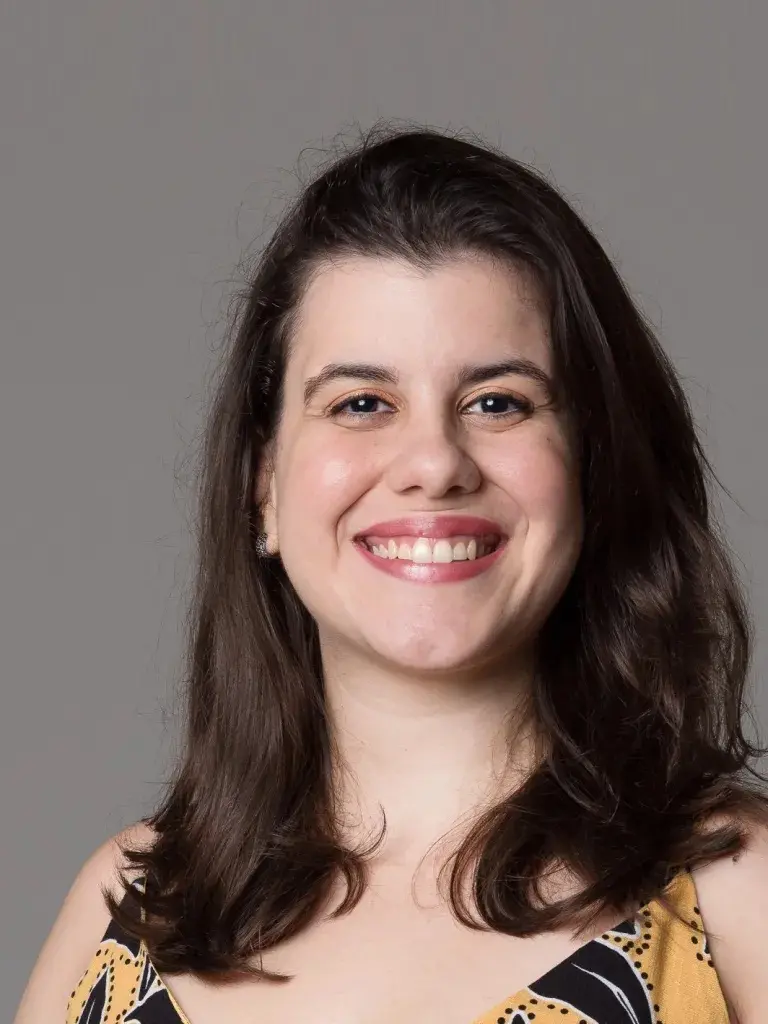 A jornalista Enoe Lopes Pontes sorri frontalmente para a câmera, exibindo cabelos castanhos ondulados e uma blusa estampada em tons de amarelo e preto. O fundo é cinza, neutro e minimalista.