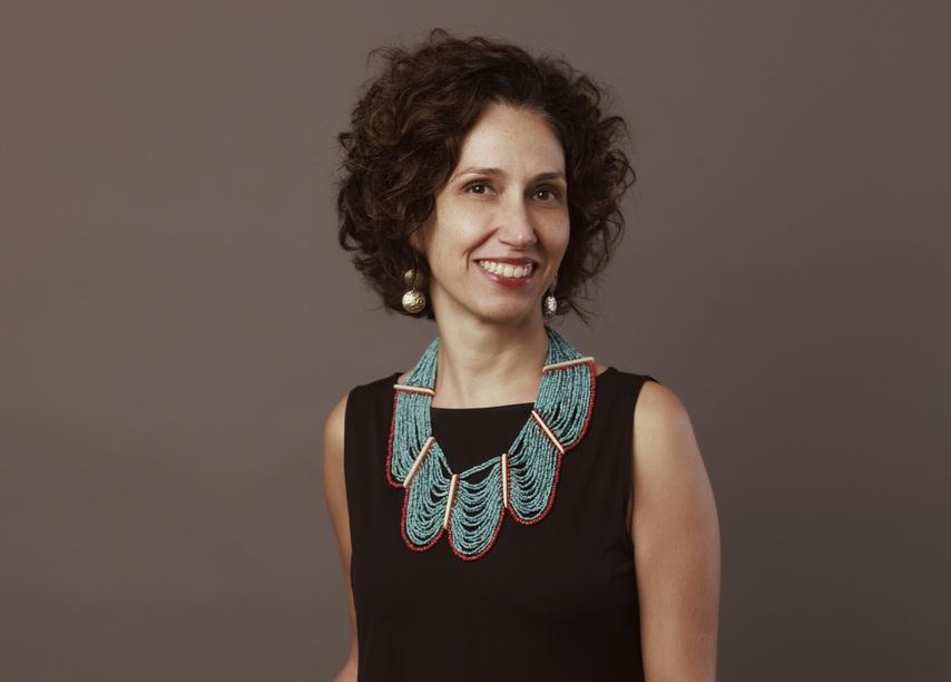 Rachel do Valle sorri usando um vestido preto e um colar de miçangas azul-turquesa de destaque, com a mão no bolso. A mulher possui cabelos curtos e cacheados, posando em um plano médio contra um fundo marrom neutro.