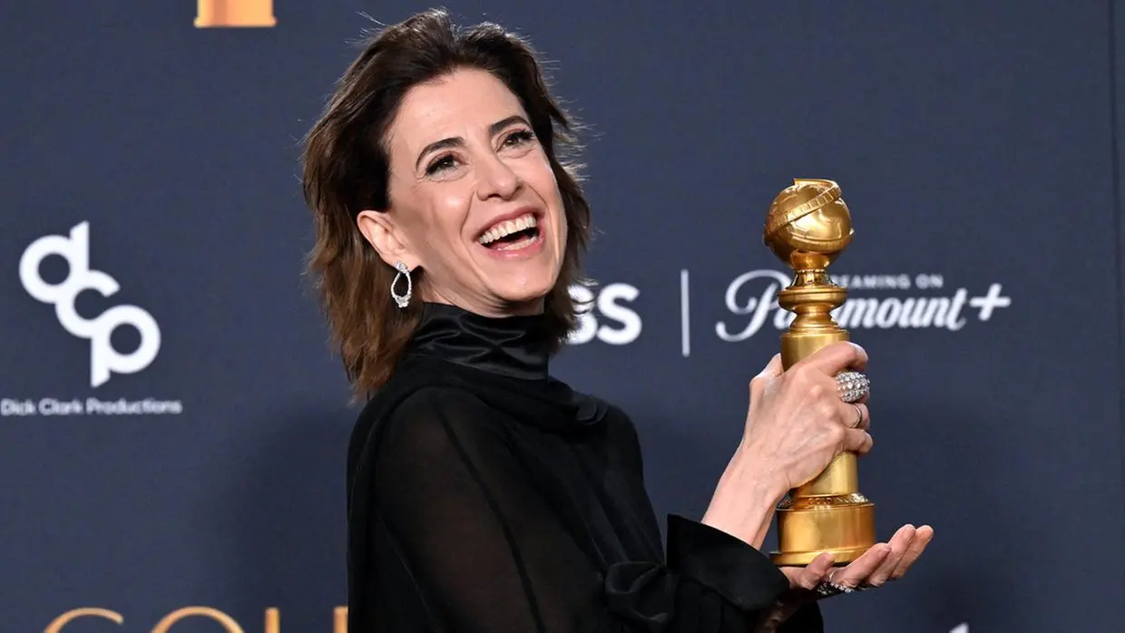 Fernanda Torres sorri com entusiasmo enquanto segura o troféu do Globo de Ouro com as duas mãos à frente de um painel de patrocinadores. Ela usa um vestido preto de gola alta e acessórios prateados, em uma composição que destaca sua expressão de celebração e vitória.