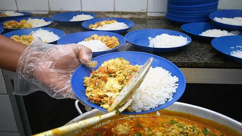 A foto mostra uma pessoa servindo um prato, com arroz e frango, em um contexto de merenda escolar no Brasil.