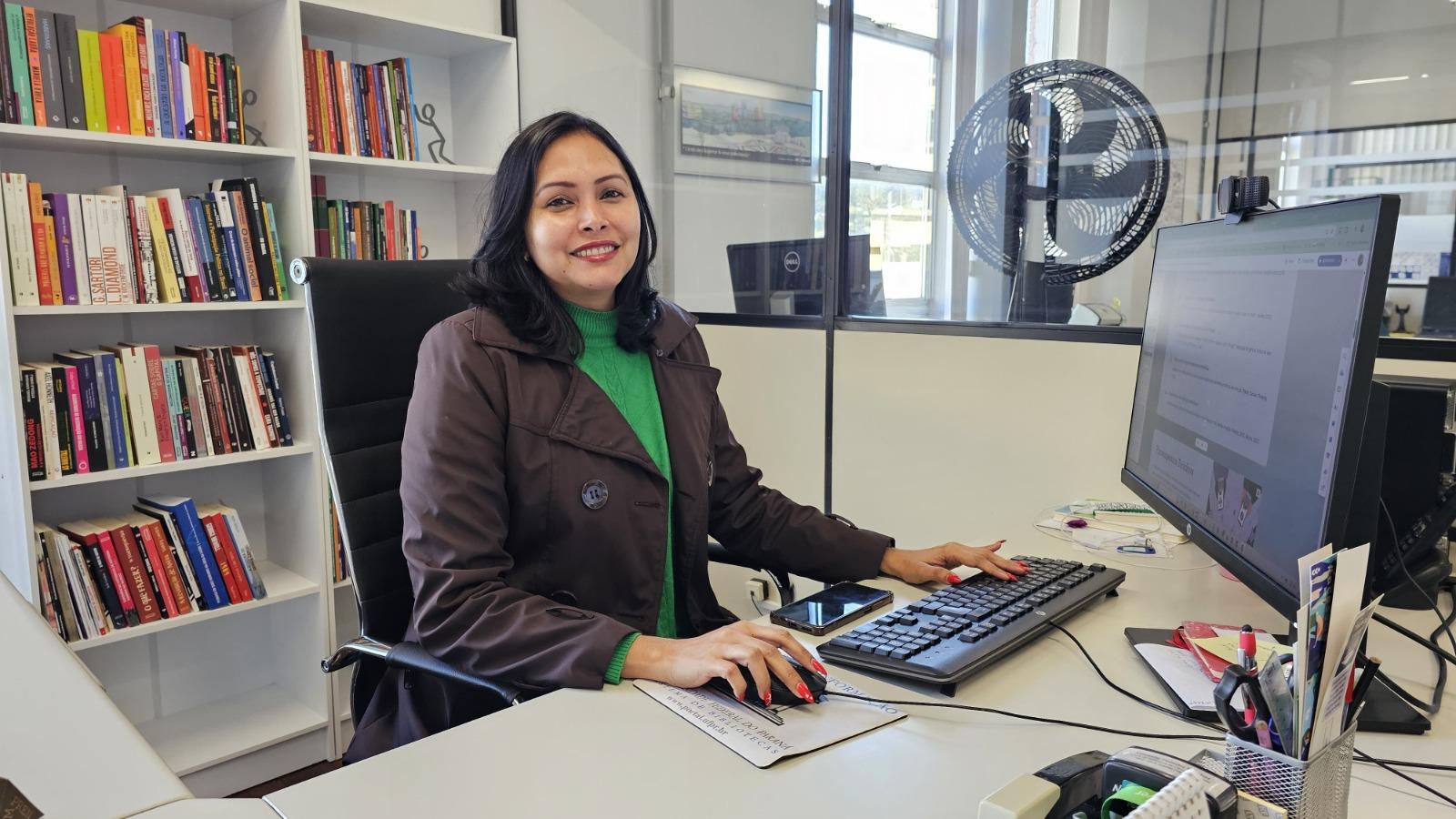 Mulher sentada à mesa de trabalho em um escritório, sorrindo para a câmera enquanto usa computador. Ao fundo, estantes com livros e uma divisória de vidro; sobre a mesa, teclado, mouse, celular e materiais de escritório.