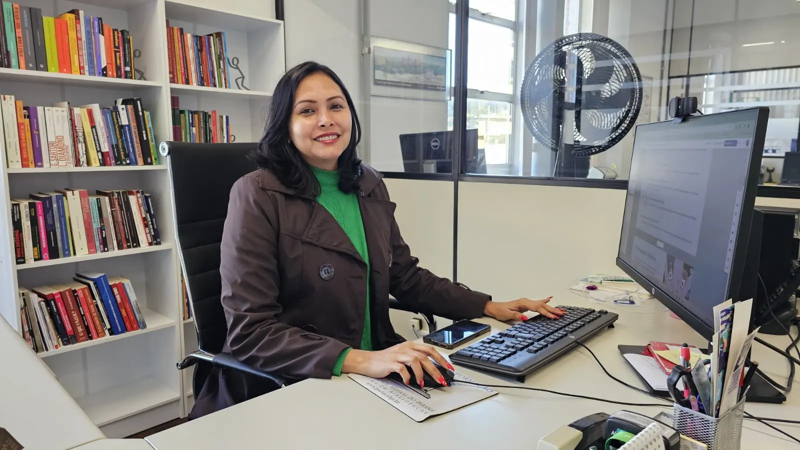 Mulher sentada à mesa de trabalho em um escritório, sorrindo para a câmera enquanto usa computador. Ao fundo, estantes com livros e uma divisória de vidro; sobre a mesa, teclado, mouse, celular e materiais de escritório.