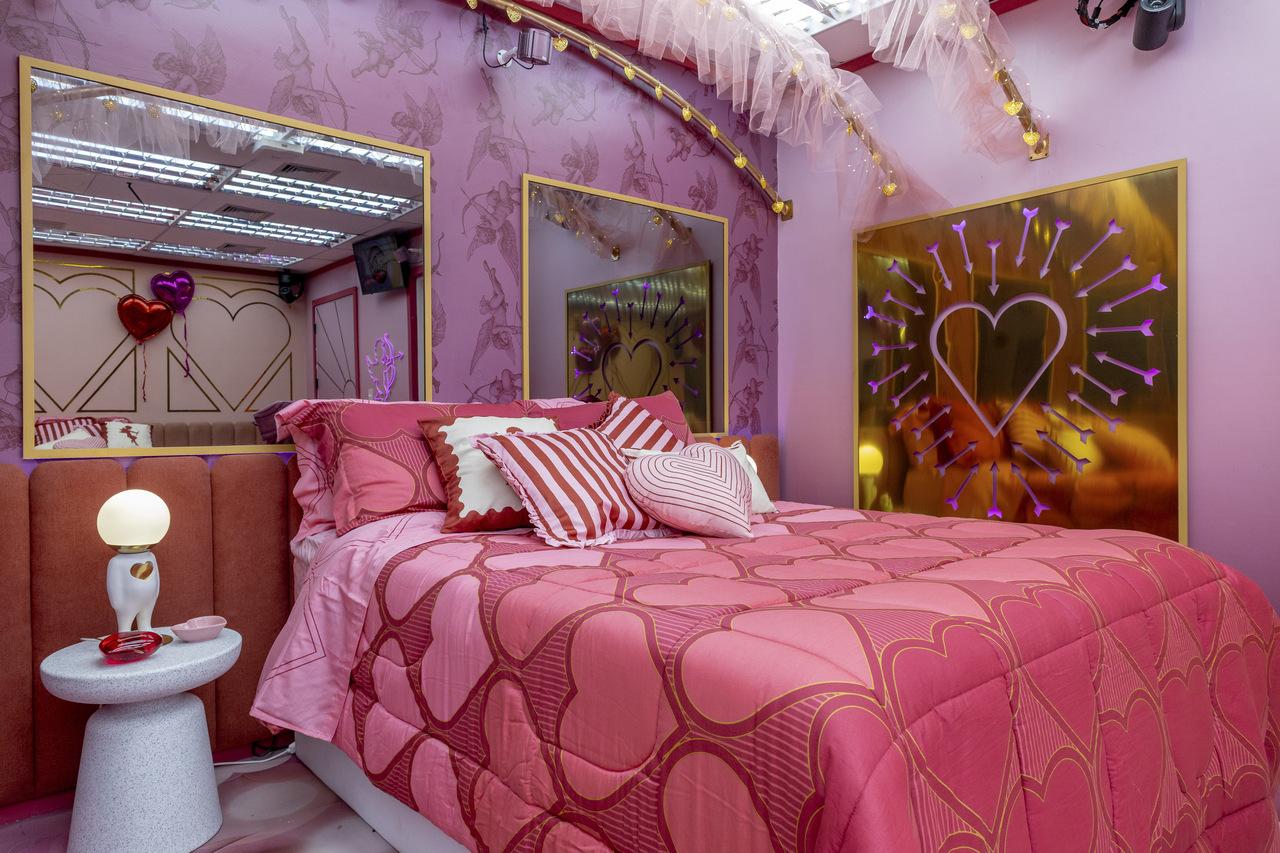 Quarto temático romântico com decoração em tons de rosa, apresentando cama central com colcha estampada, almofadas coordenadas e iluminação aconchegante. O ambiente conta com painéis decorativos dourados com corações iluminados, parede com textura artística, espelhos com molduras elegantes e elementos cenográficos delicados, criando um espaço sofisticado, intimista e visualmente impactante.