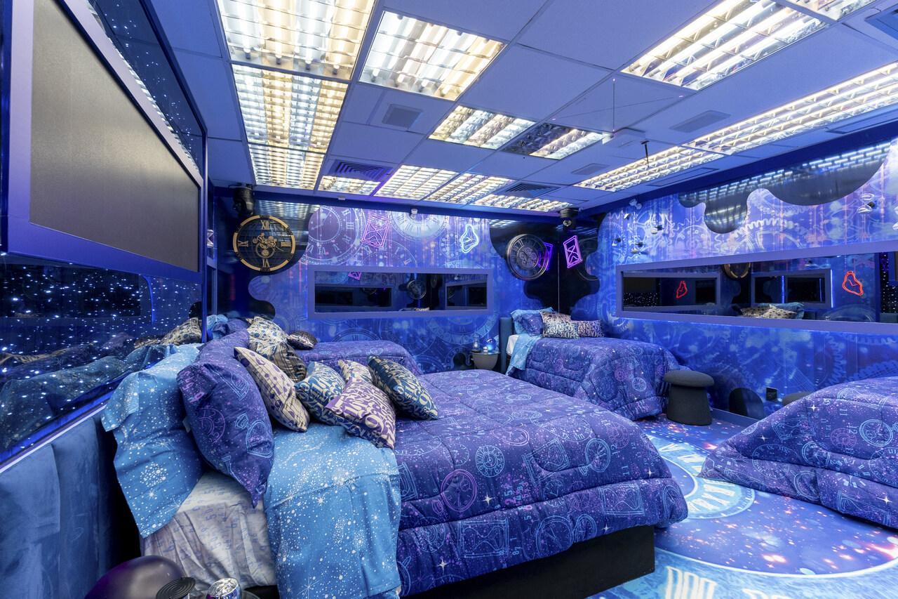 Quarto temático futurista com decoração em tons de azul e roxo, apresentando camas amplas com edredons estampados de constelações, símbolos astronômicos e padrões cósmicos. O ambiente conta com paredes e piso ilustrados com elementos espaciais, iluminação cênica no teto, espelhos laterais integrados, almofadas decorativas coordenadas e detalhes luminosos que criam uma atmosfera imersiva, moderna e visualmente impactante, típica de um cenário de reality show ou espaço cenográfico de alto padrão.