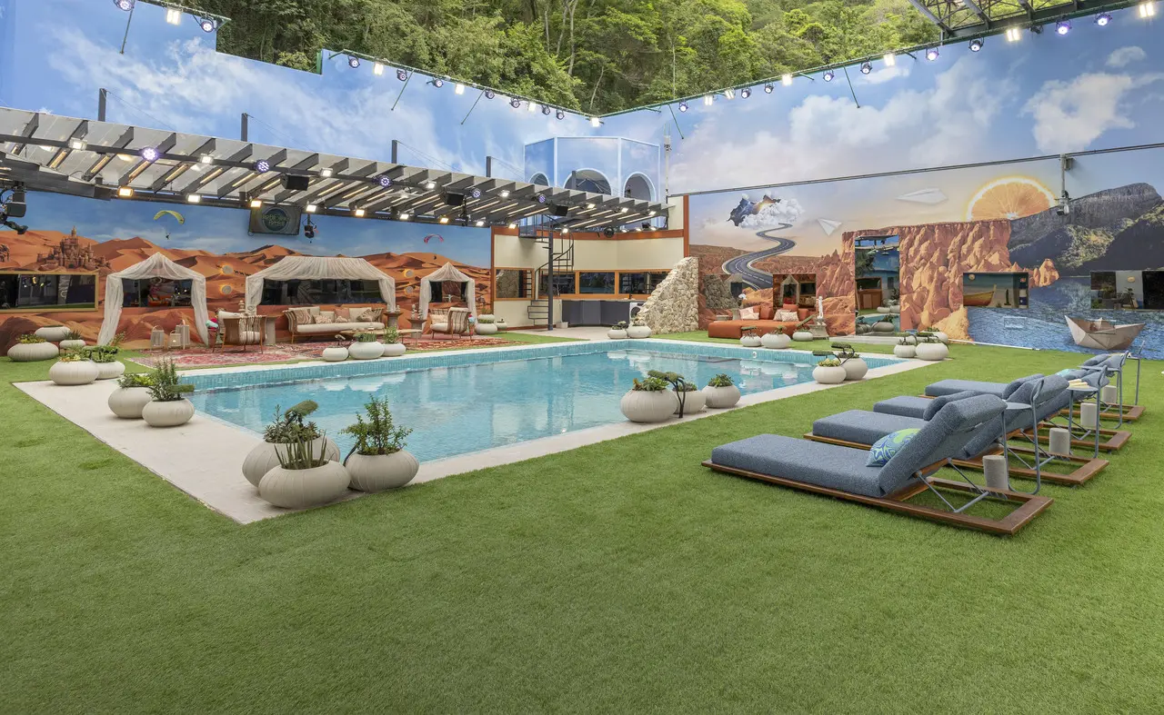 Área externa ampla com piscina retangular central, cercada por gramado bem cuidado, espreguiçadeiras acolchoadas e vasos decorativos com plantas. O espaço conta com lounges cobertos, estruturas com pergolado, iluminação cênica suspensa e paredes ilustradas com paisagens em estilo desértico e temático. O ambiente combina design contemporâneo, conforto e cenário cenográfico, criando uma atmosfera de lazer sofisticada típica de um espaço residencial de alto padrão ou cenário de reality show.
