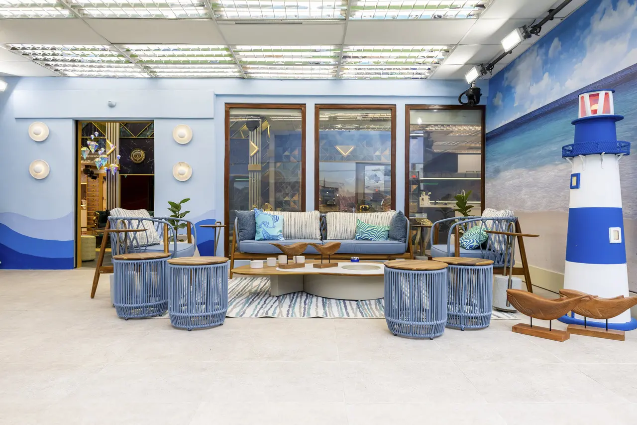 Varanda cenográfica em ambiente interno que remete a um espaço praiano, com sofás, poltronas e mesas em tons de azul e madeira dispostos diante de fachadas envidraçadas. O cenário inclui painel ilustrado com paisagem litorânea e farol decorativo, além de iluminação profissional no teto, evidenciando um espaço preparado para gravações e uso televisivo em contexto de reality show ou produção audiovisual.