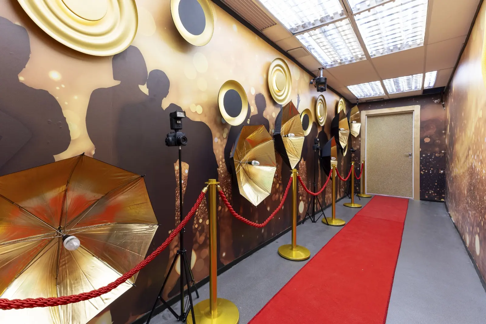 foto do caminho do quarto do lider do BBB 26, com tapete vermelho e rebatedores dourados com luzes de flash piscando, silhuetas de fotógrafos e câmeras em tripé.