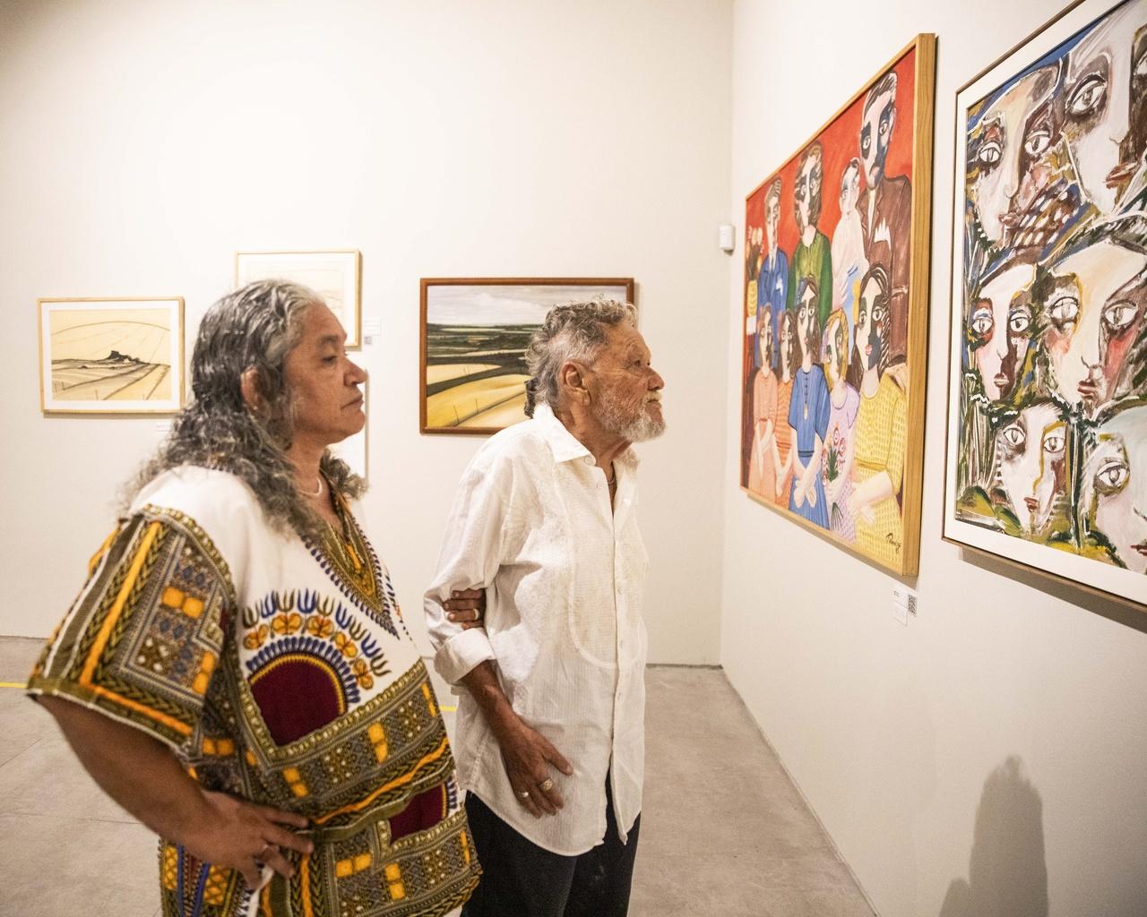 O curador Gerson Ipirajá, vestido de túnica estampada, e o artista Zé Tarcísio, vestindo camisa branca de manga longa, observam quadros coloridos com figuras humanas em uma galeria de arte com paredes brancas.