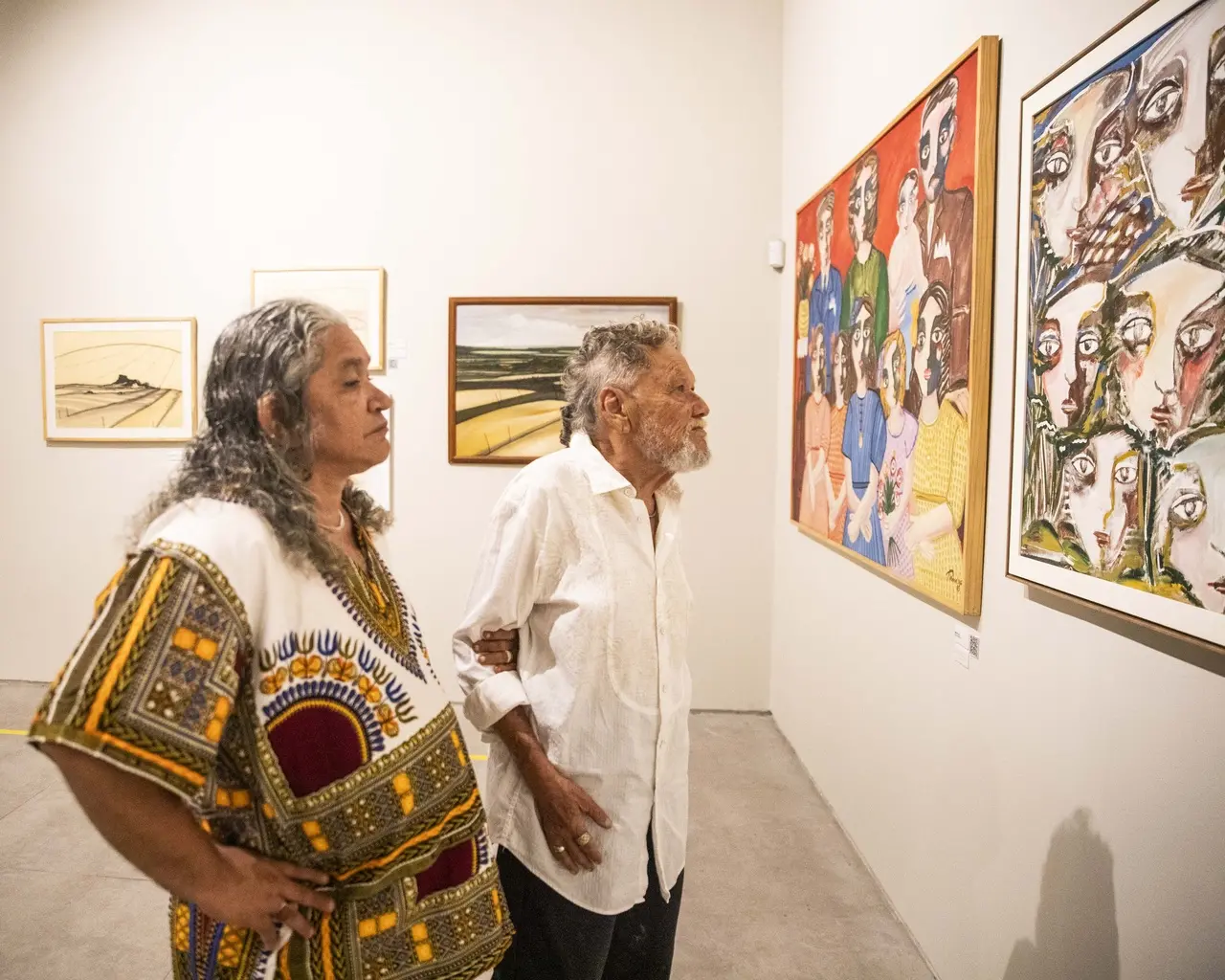 O curador Gerson Ipirajá, vestido de túnica estampada, e o artista Zé Tarcísio, vestindo camisa branca de manga longa, observam quadros coloridos com figuras humanas em uma galeria de arte com paredes brancas.