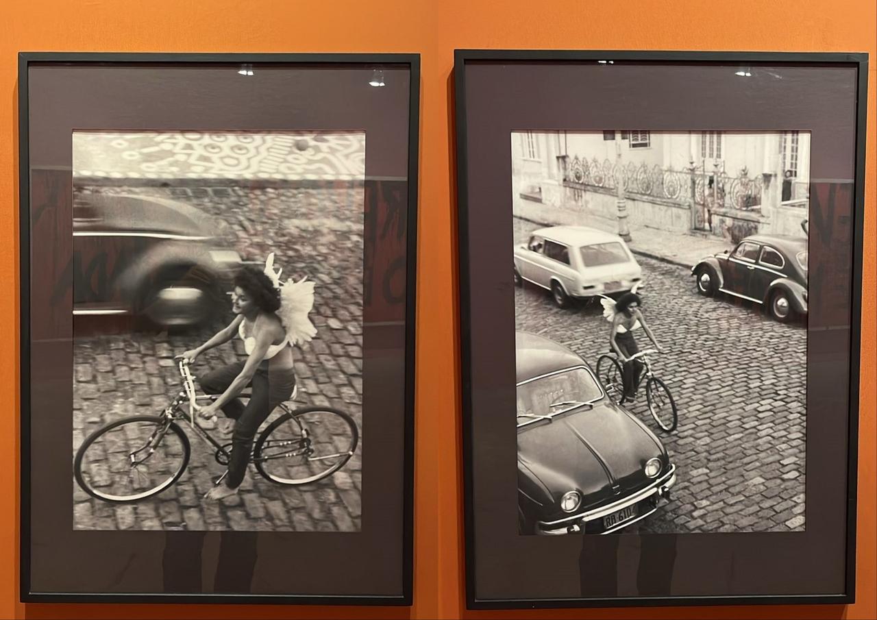 Montagem de duas obras de arte em preto e branco que mostram uma mulher andando de bicicleta trajando asas de anjo. Ambas estão colocados em uma parede laranja.